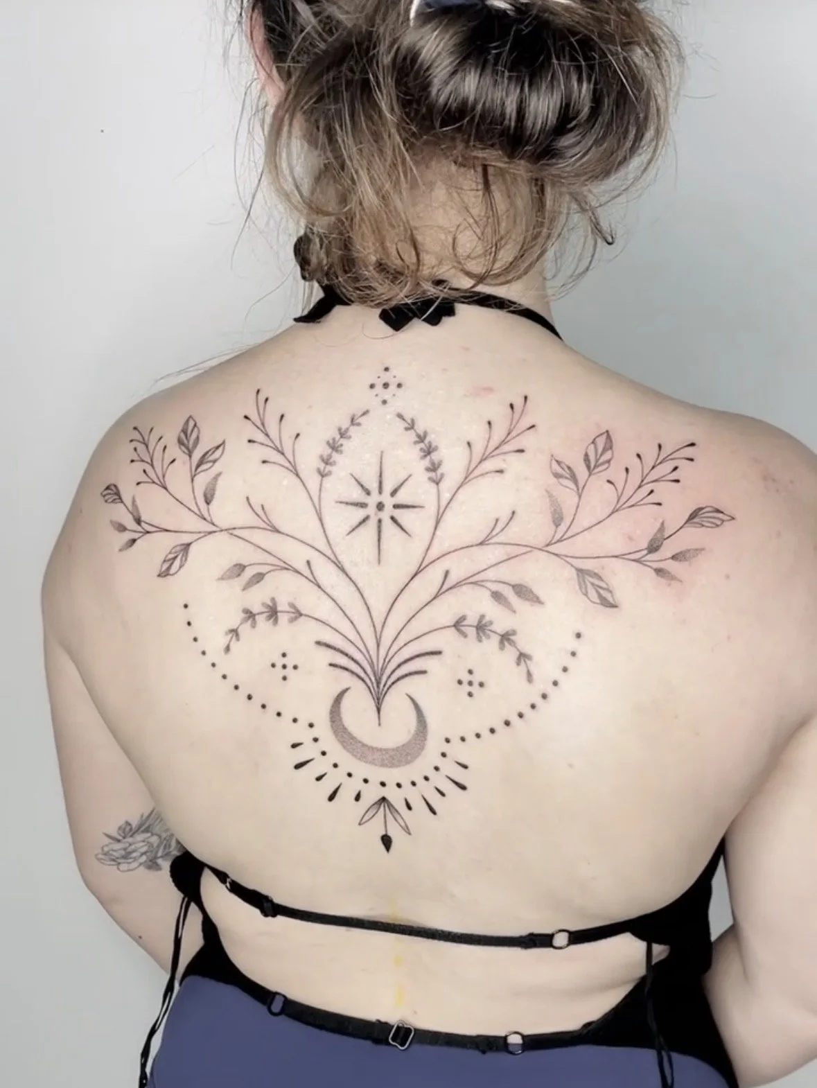 A person's back with a black ink tattoo featuring a crescent moon, crescent stars, and floral designs arranged symmetrically.