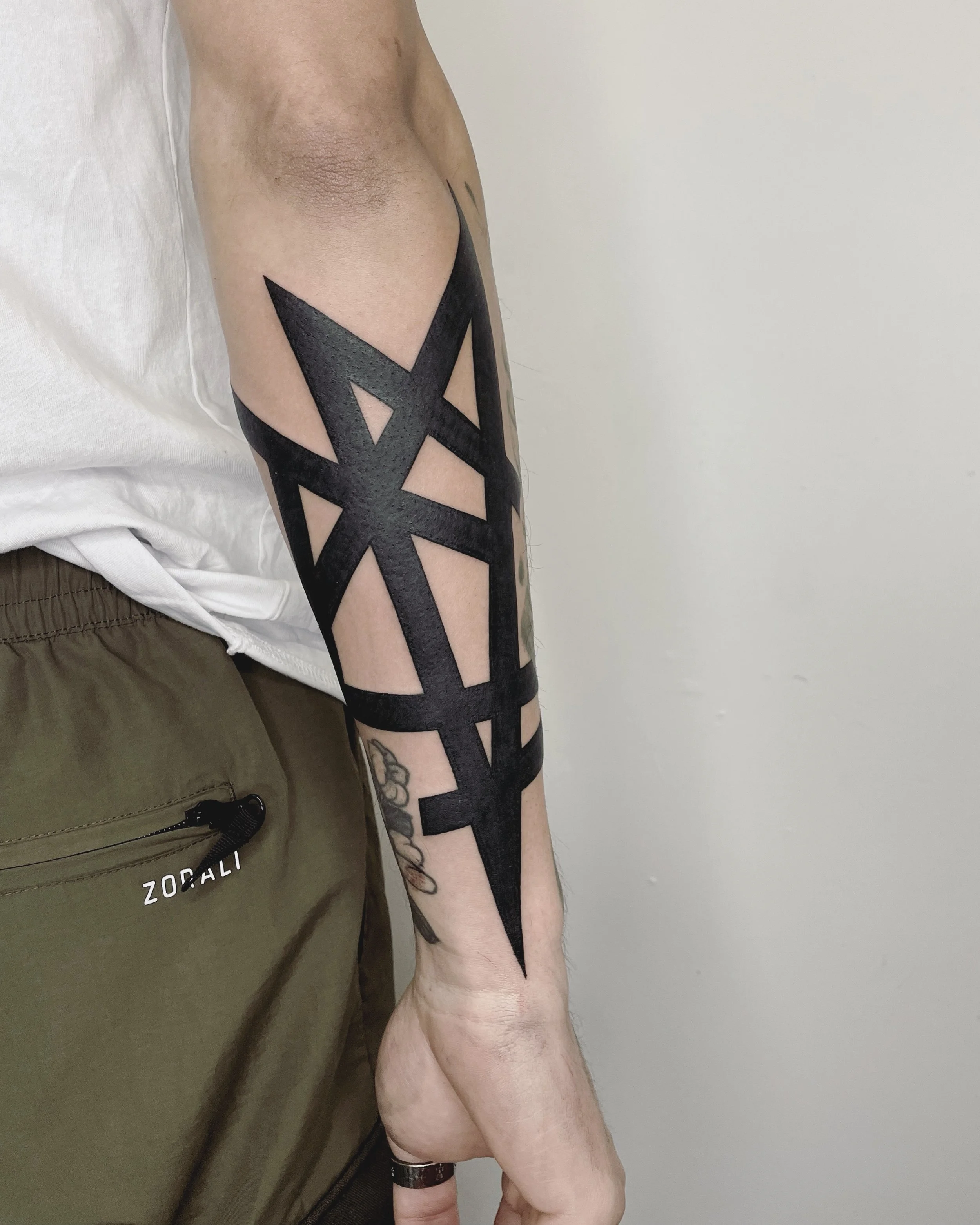 Close-up view of a person's arm with tribal-style black tattoos, next to the torso dressed in a white shirt and khaki shorts. The tattoos feature bold geometric and symmetrical patterns extending from shoulder to forearm.