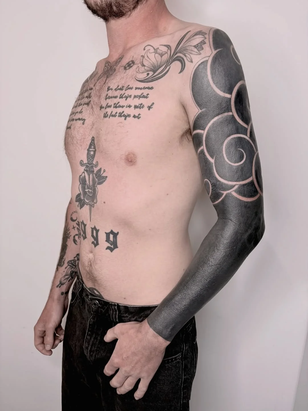 A tattooed man with a full sleeve of black tribal and floral designs on his right arm, a rose with a dagger tattoo on his torso below the chest, and a script tattoo on his chest that reads, 'we didn't love someone have they perfect we love them in sp