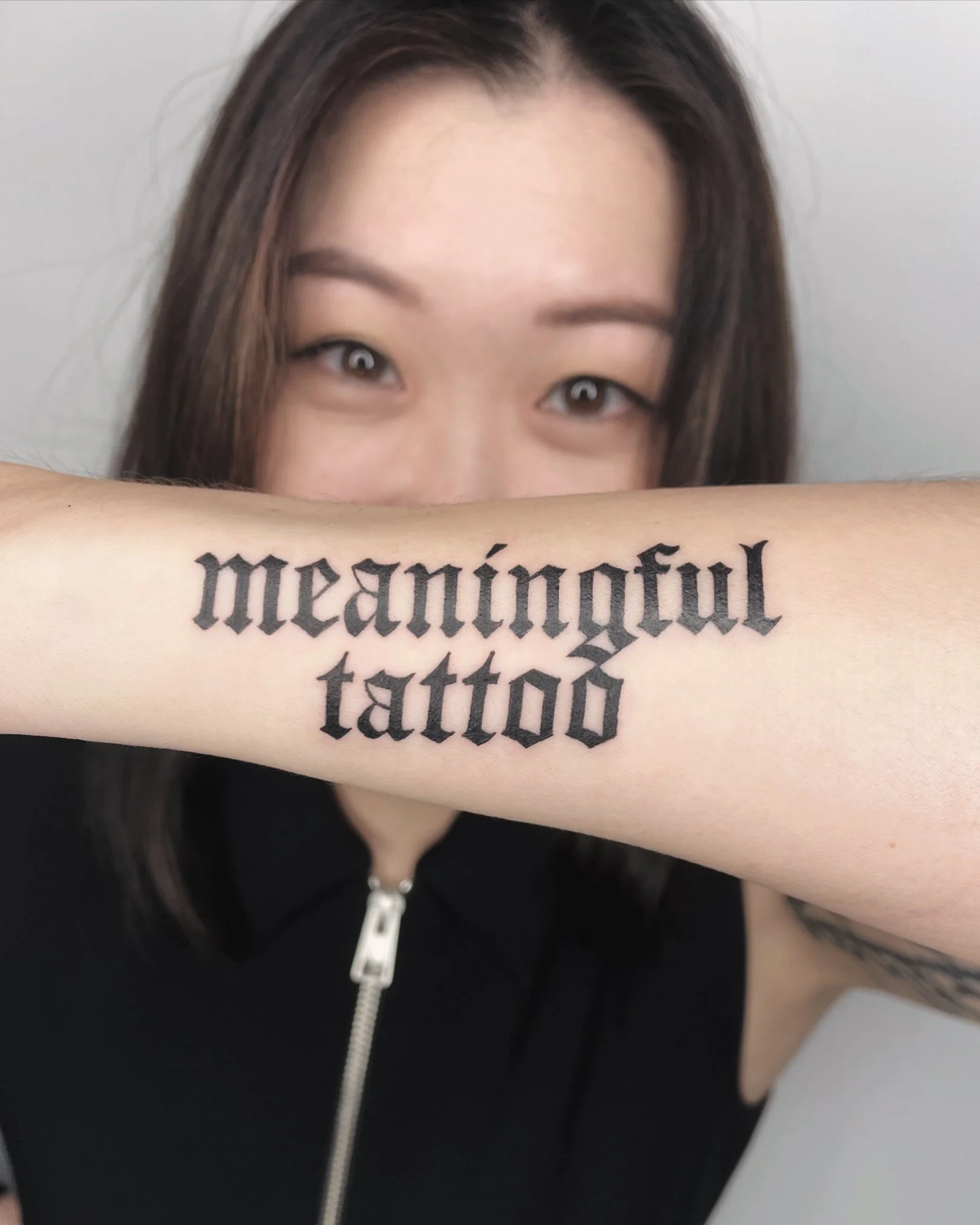 A woman with dark hair and brown eyes hiding her face behind her forearm, which has a large tattoo of Gothic-style black text.