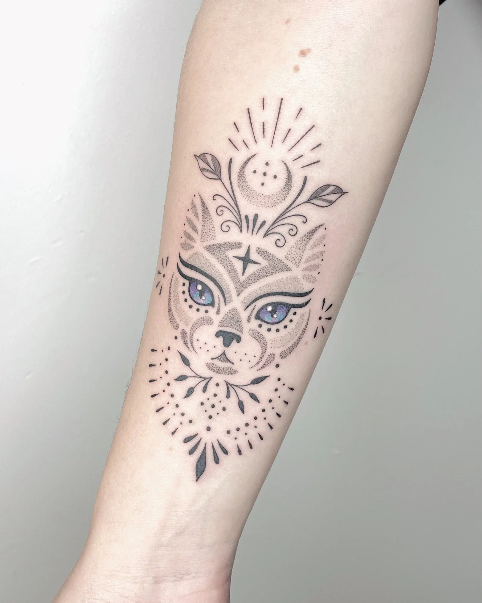 Tattoo of a cat's face with purple and blue eyes, surrounding it are decorative elements like leaves, stars, and sun rays on a person's forearm.