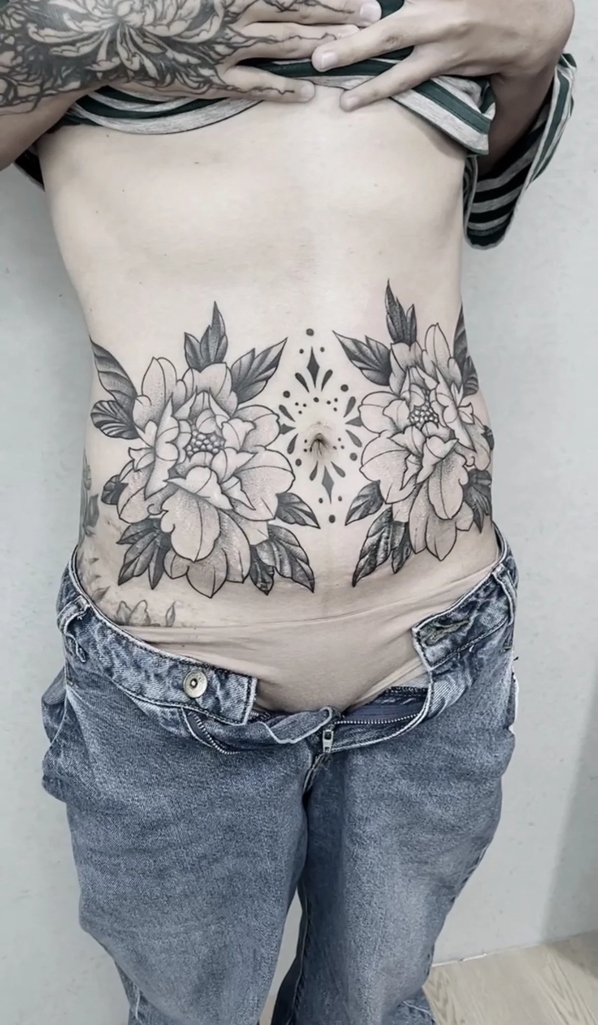 A person with tattoos on their chest and arms lifting their shirt to reveal a floral tattoo on their torso, wearing jeans.