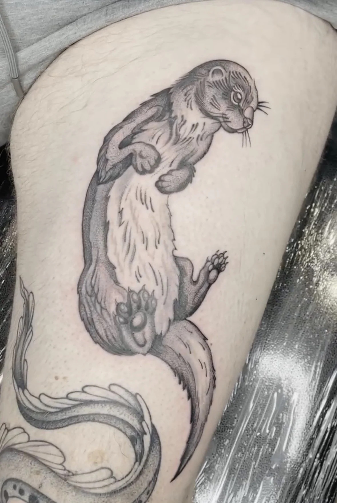 A black and grey tattoo of a mythical creature with the body of a snake and the head of a feline, depicted in a traditional tattoo style.