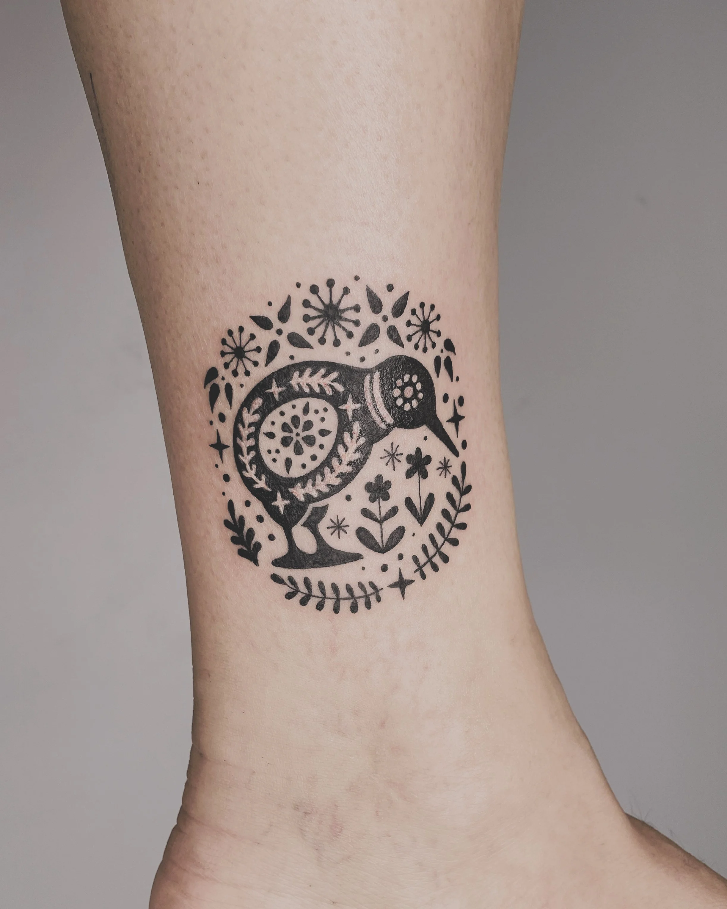 Tattoo of a bird surrounded by floral and decorative patterns on a person's ankle.