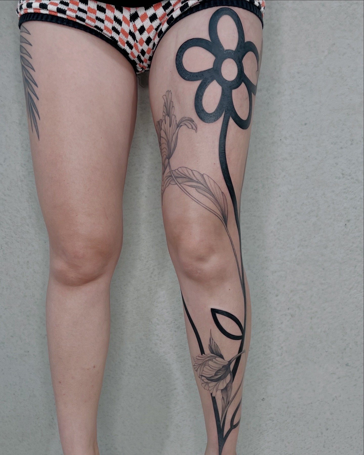 Person with tattoos on their upper thigh and lower leg, wearing patterned shorts, against a plain wall.