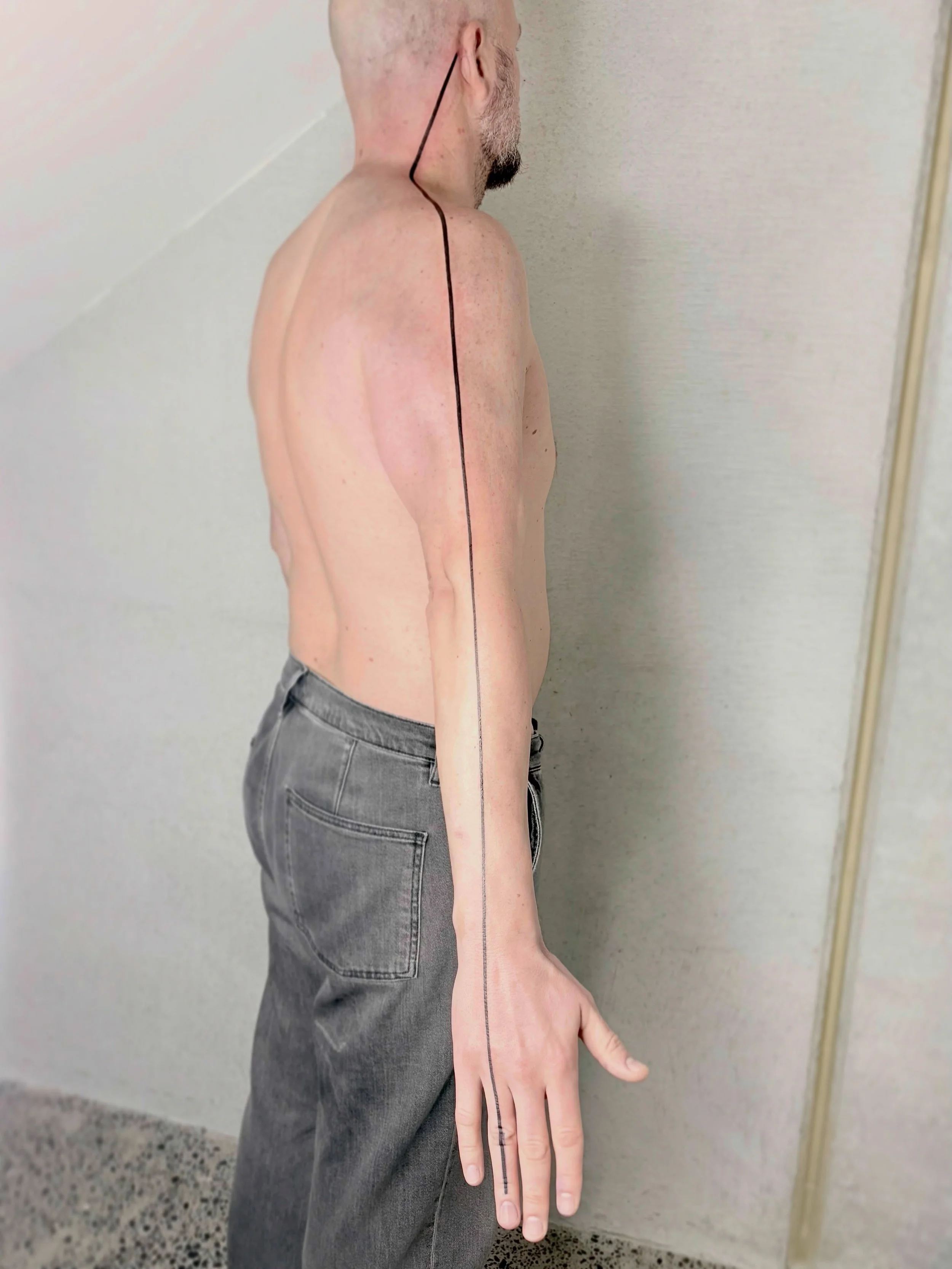 A man standing against a wall with a black line drawn from his ear down to his little finger, indicating the line of the body's natural posture.