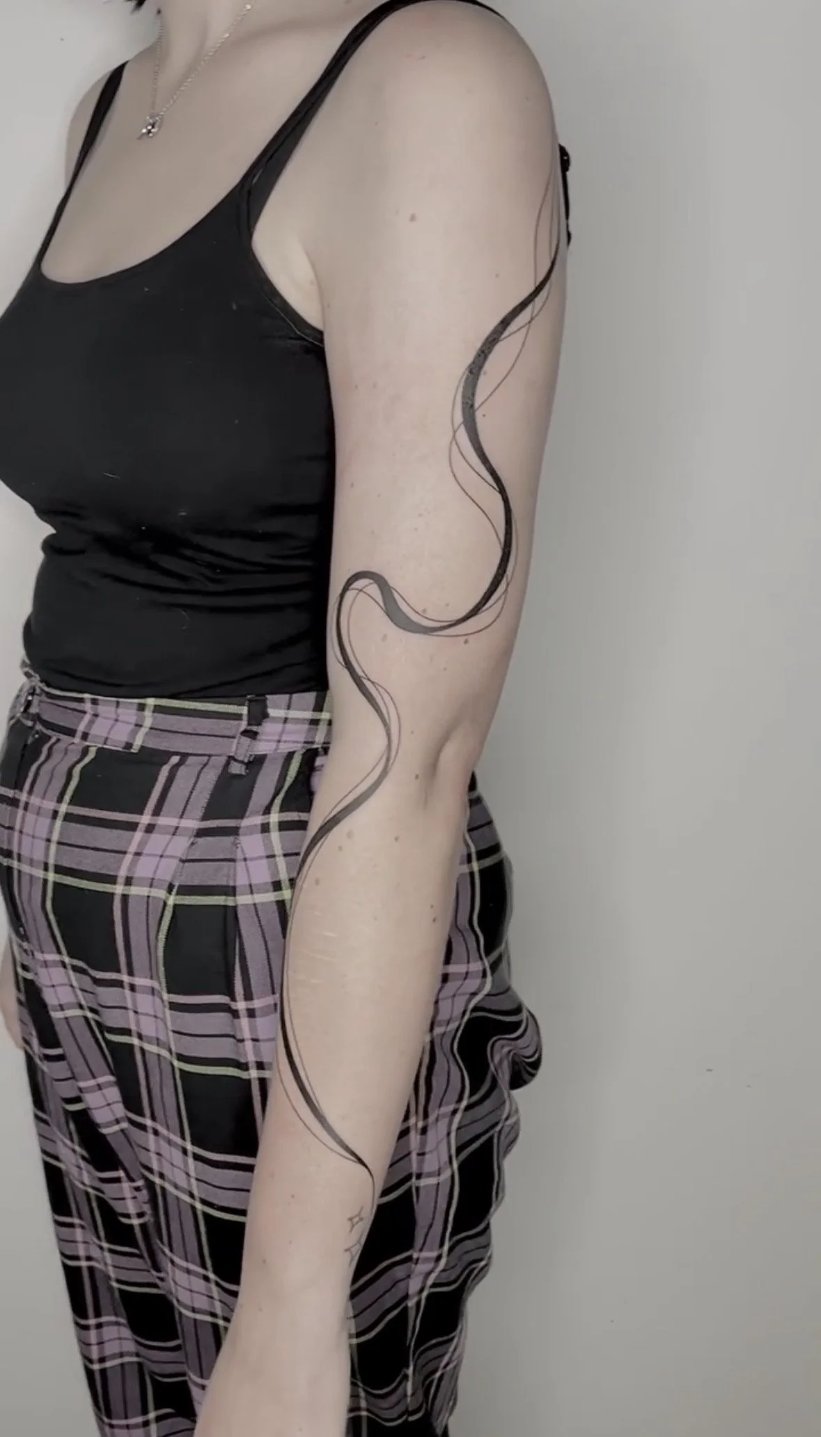 A person showing a tattoo of a long, wavy black line running down their arm and side, dressed in a black tank top and plaid pants.