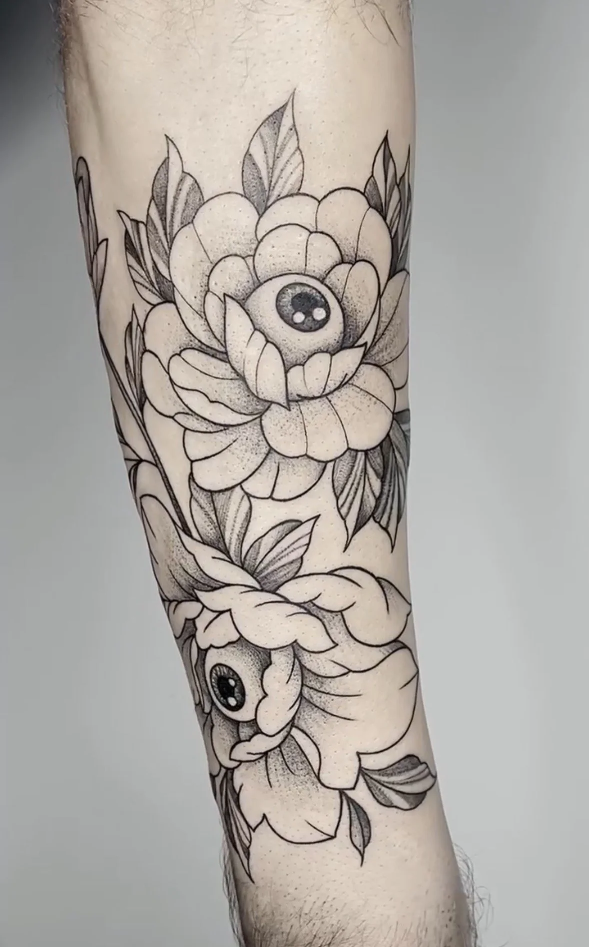 Black and gray floral tattoo on a person's arm featuring a large flower with an eye in the center, surrounded by leaves.