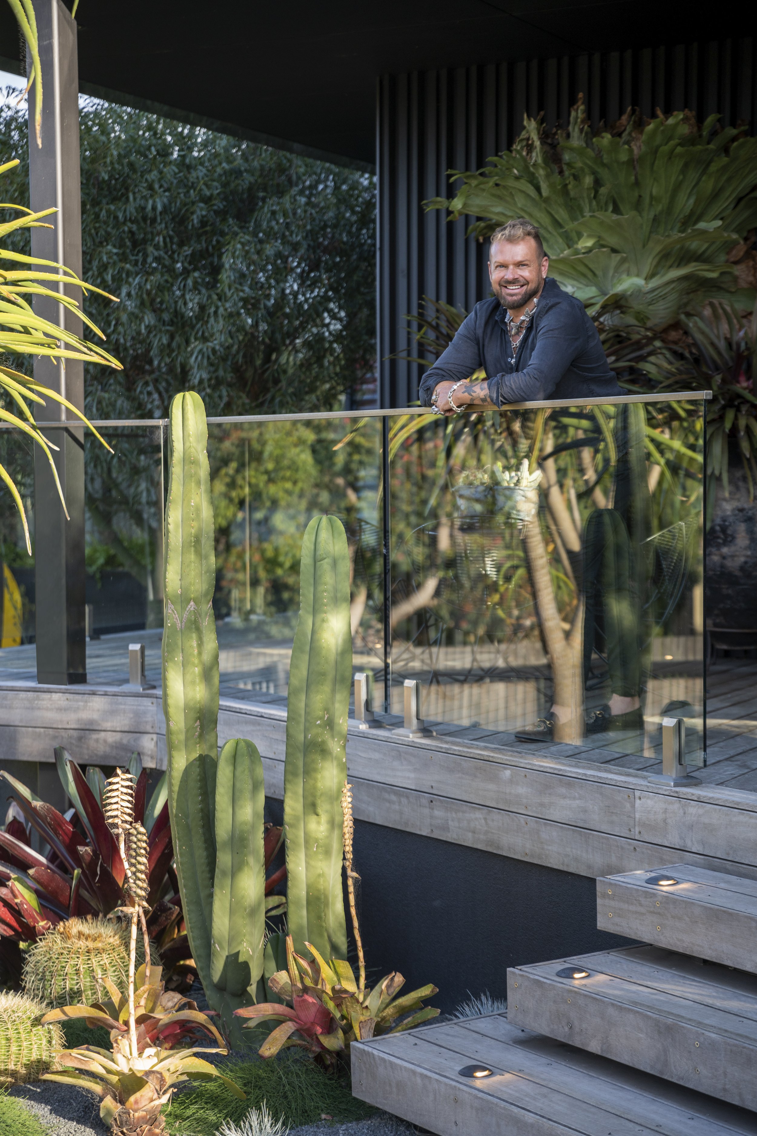 A person leaning on a glass railing of a house's deck, surrounded by cacti and lush greenery.