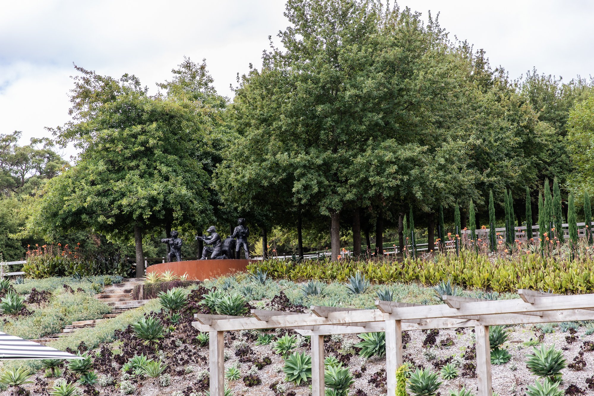 Outdoor garden scene with various plants, trees, and a group of dark sculptures on a stone platform in the background.