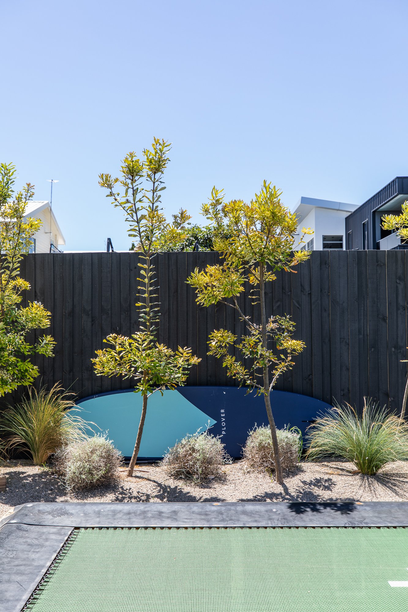 Outdoor scene with green trampoline, small trees, shrubs, and a surfboard against a wooden fence under clear blue sky.