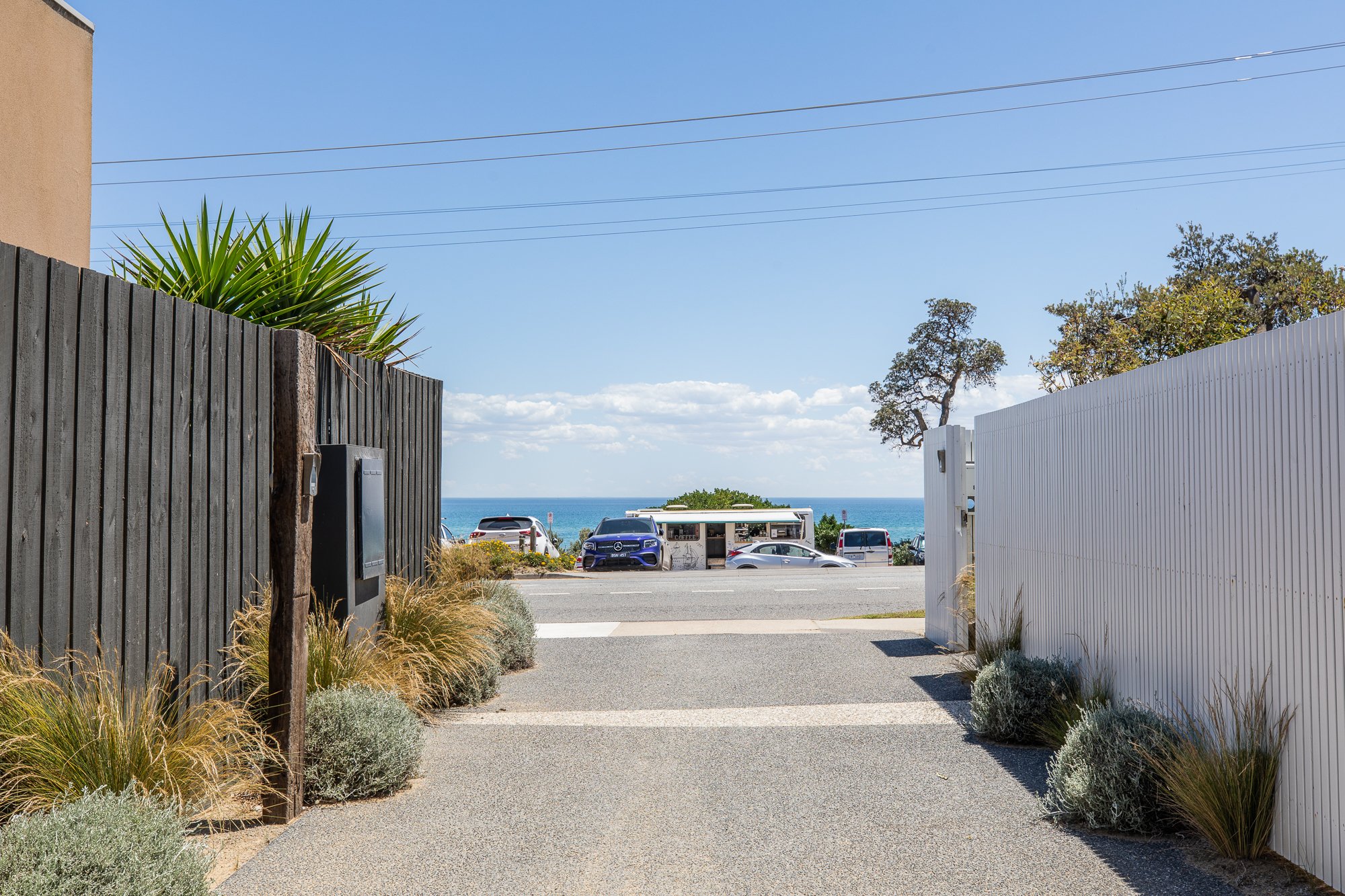 Pathway leading to a street with parked cars, beach view in the background, flanked by wooden fences and greenery, under a clear blue sky.