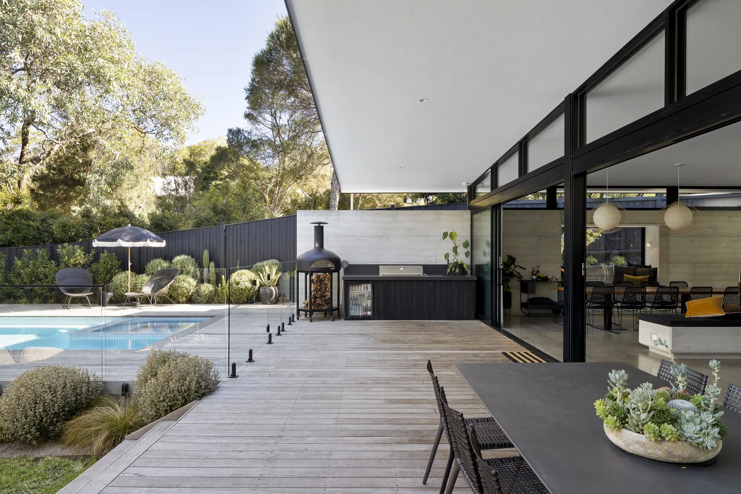 Modern outdoor patio with wooden decking, glass fence around a swimming pool, outdoor kitchen with a wood-fired oven, dining area with table and chairs, surrounded by plants and trees.