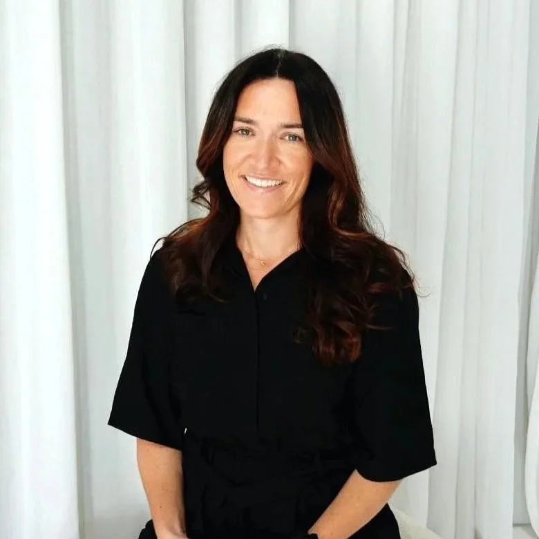 A woman with long wavy brown hair, wearing a black shirt, smiling in front of white curtains.