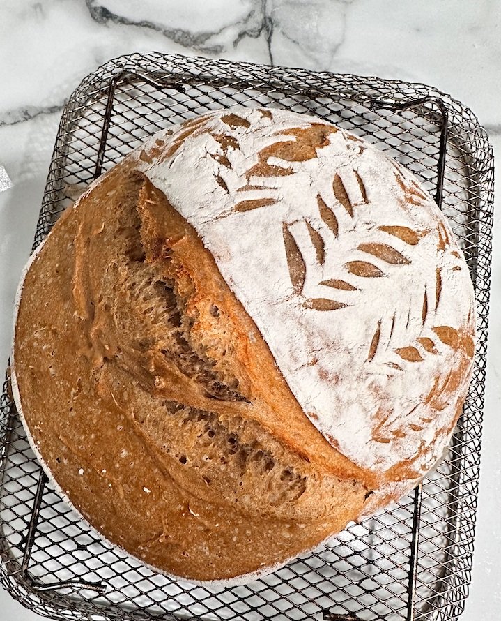 Freshly baked round loaf of sourdough bread with decorative scoring on top, placed on a wire cooling rack.