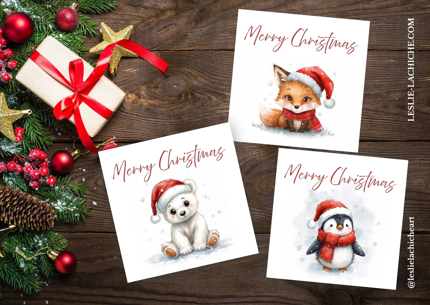 Three Christmas cards with cute animal illustrations wearing Santa hats, on a wooden surface with holiday decorations including a small gift, pine branches, red ornaments, and gold star ornaments.