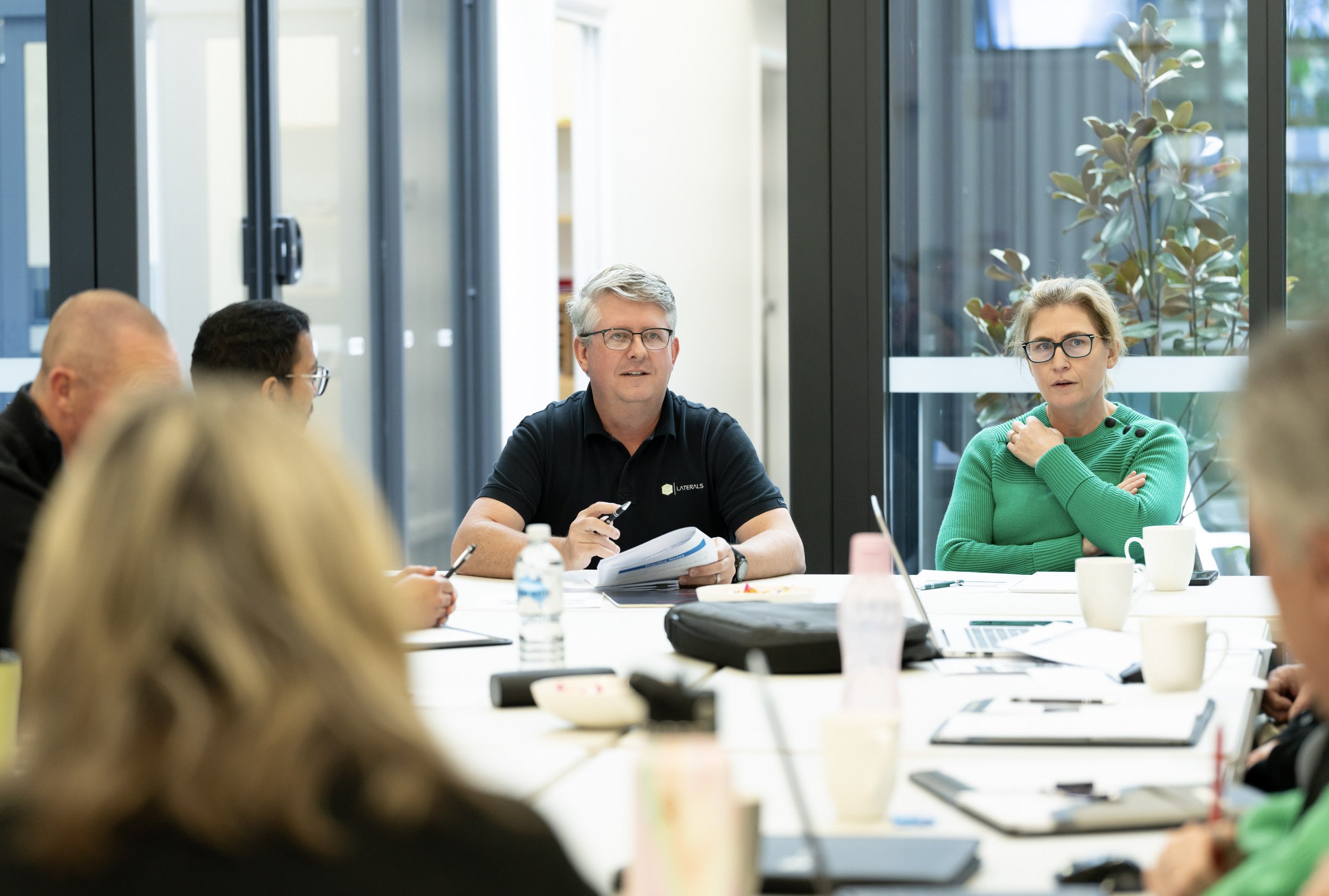 A group of people in a meeting room with large windows, sitting around a table with laptops, documents, cups, and bottled water, engaged in a discussion.