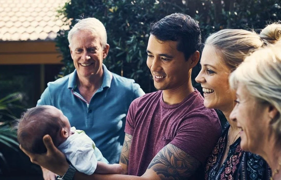A young man holding a newborn baby, surrounded by smiling older adults in a backyard setting.