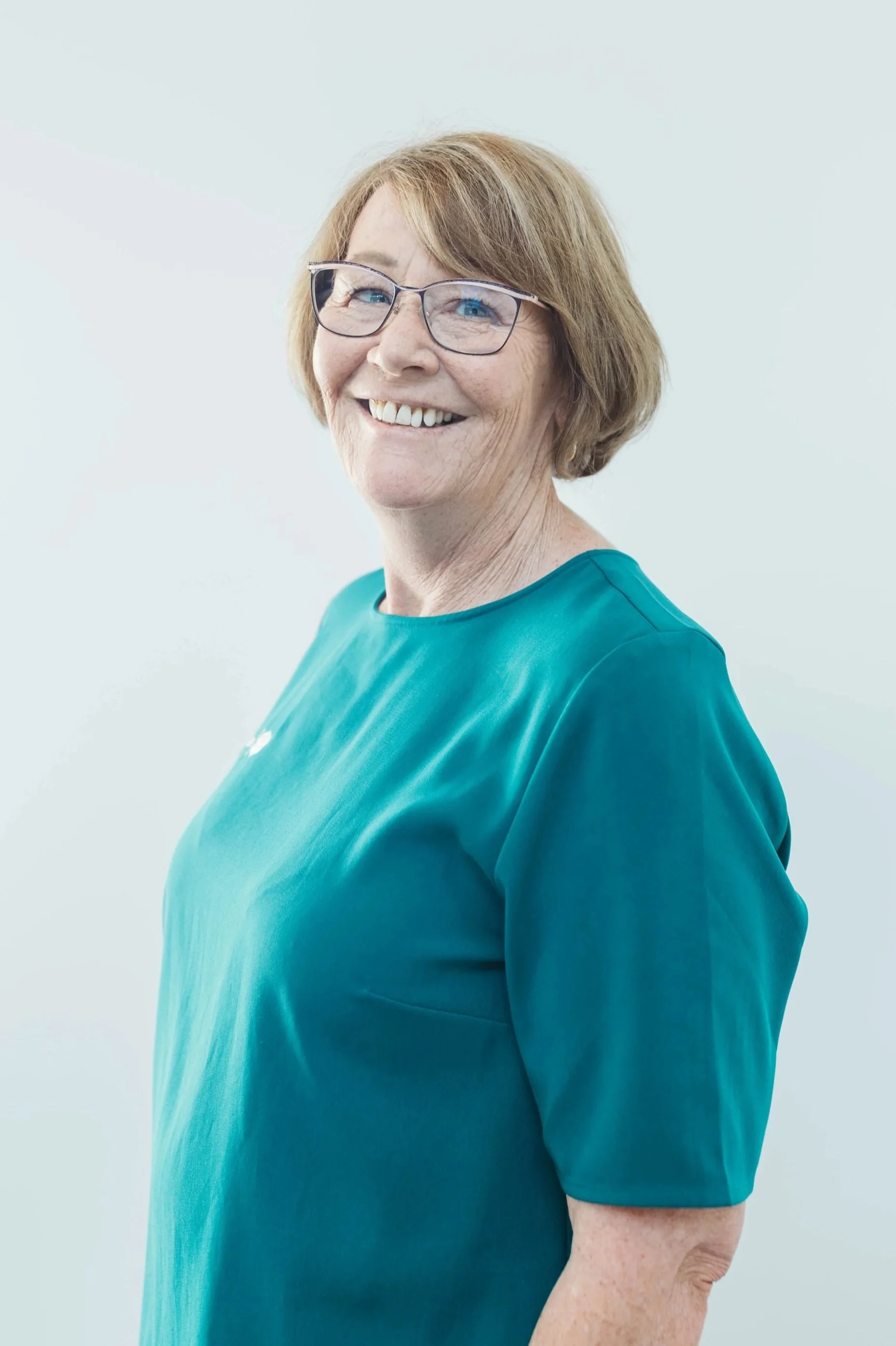 A smiling woman with glasses and short, light brown hair, wearing a teal top, standing against a plain light background.