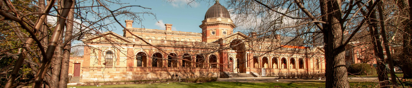 Historic building with a dome and arched windows, framed by trees in the foreground, under a partly cloudy sky.