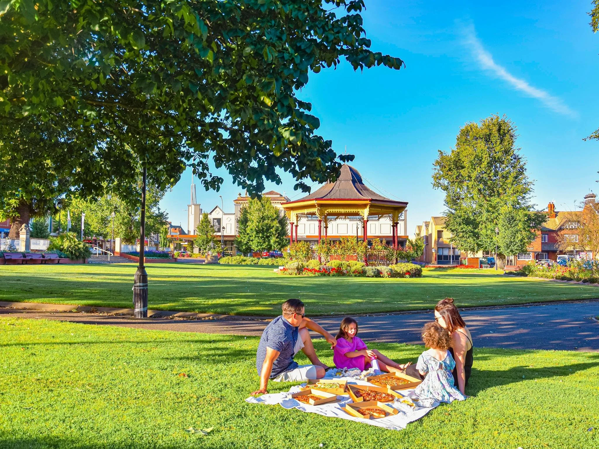 A family of four having a picnic on a blanket with pizza in a park on a sunny day, with a pavilion, trees, and houses in the background.