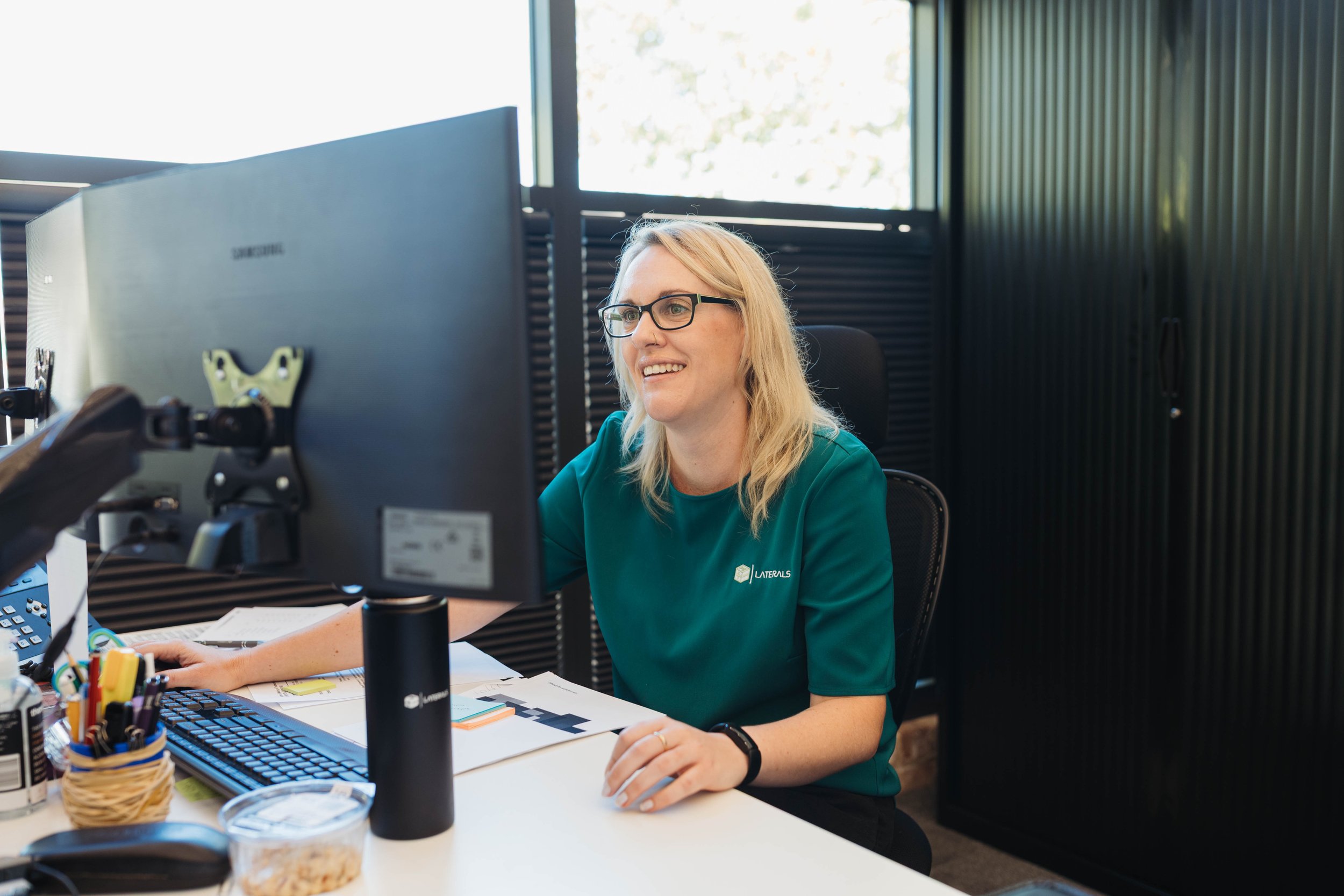 A woman with blonde hair, glasses, and a teal shirt sitting at a desk working on a computer in an office with large windows and black walls.