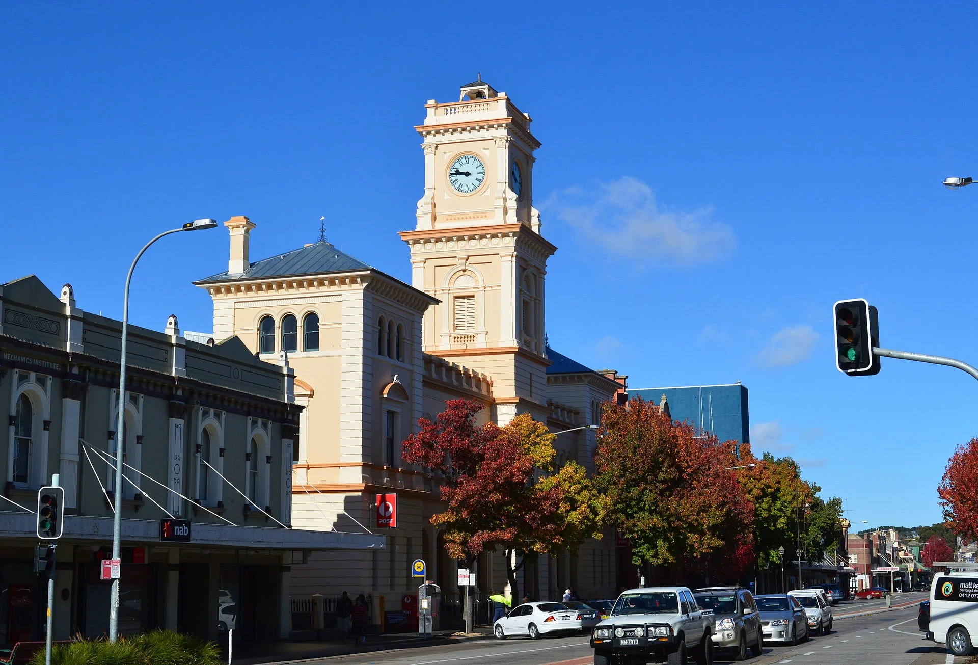A city street scene with a historic building featuring a clock tower, with cars parked along the street, trees with fall foliage, and a traffic light showing red.
