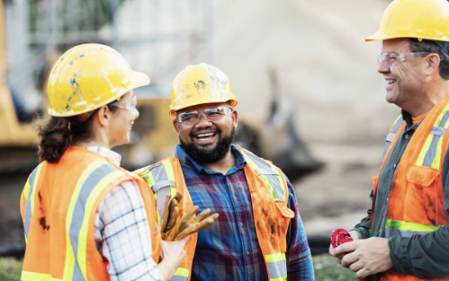 Three construction workers wearing yellow safety helmets and orange vests, smiling and talking at a construction site.