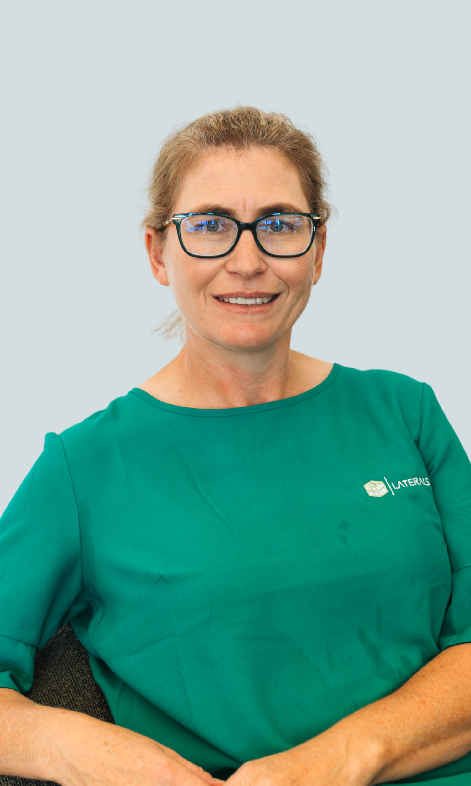 A woman wearing glasses and a green shirt with the 'Latern' logo, sitting and smiling in front of a plain light blue background.