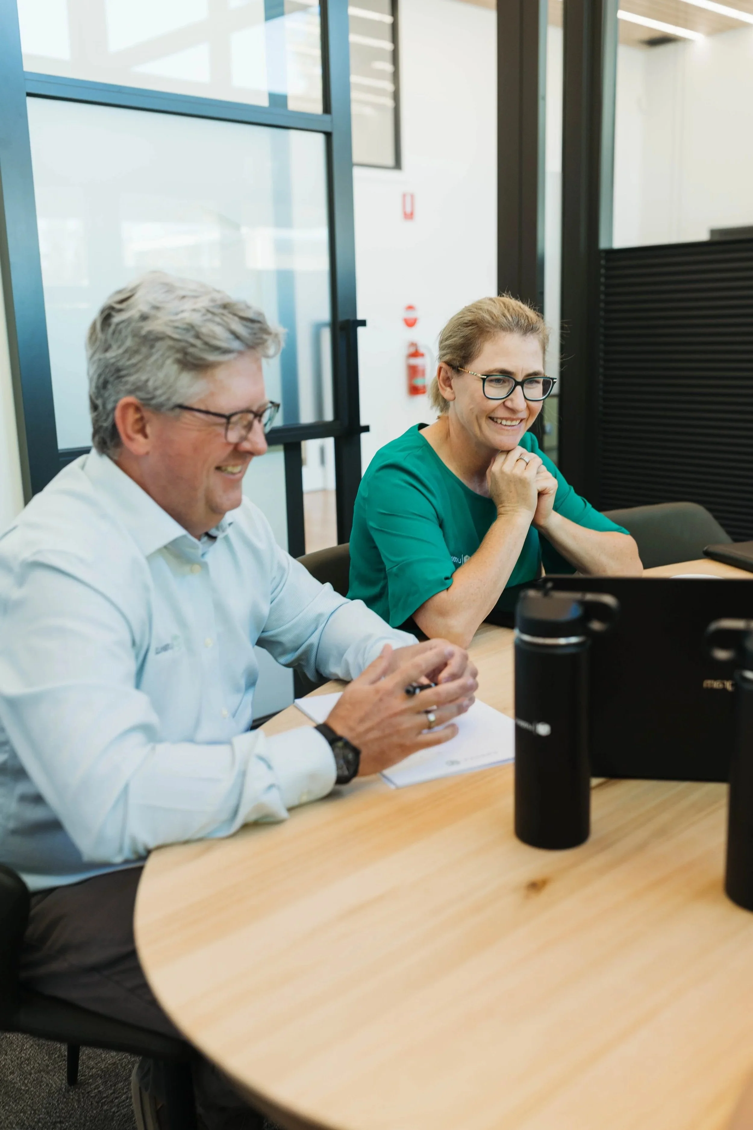 Two professionals, a man and a woman, sitting at a conference table in an office, smiling and looking at a computer screen.