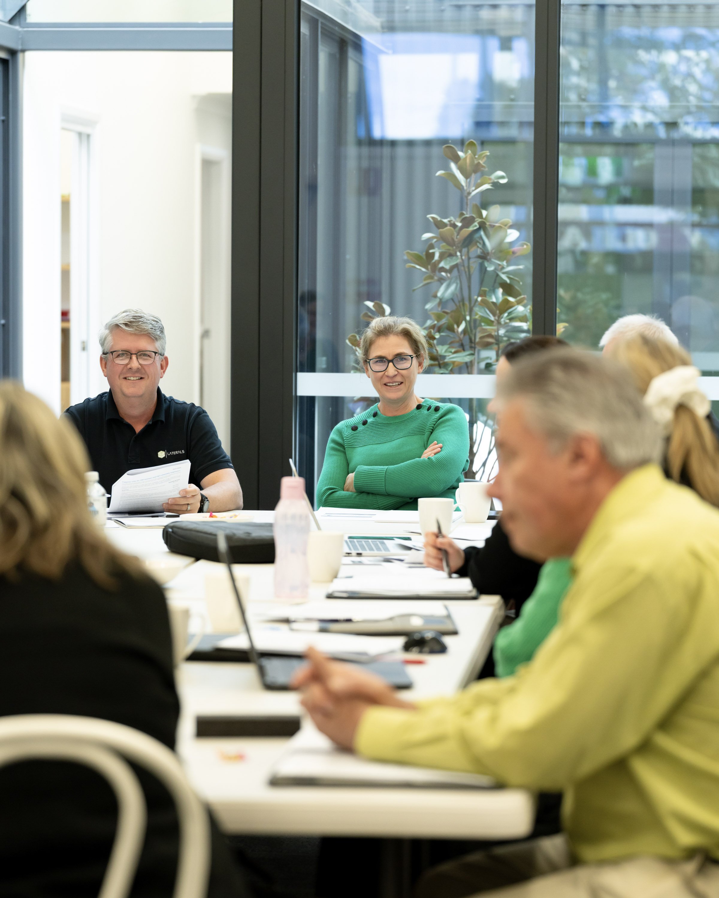 People in a meeting room sitting around a table, engaged in discussion, with a large window showing plants outside.
