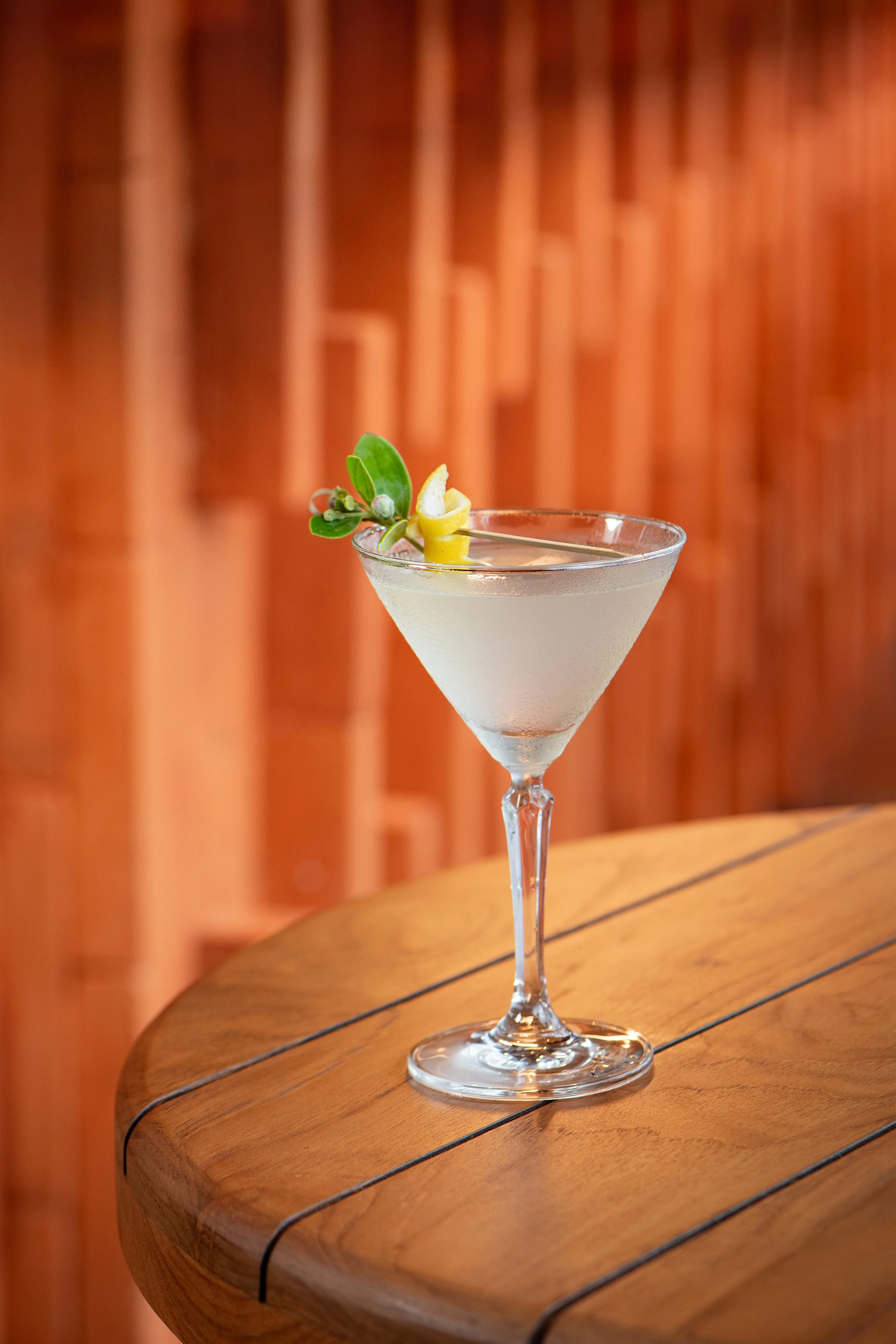 A classic cocktail served in a martini glass, garnished with a lemon twist and a sprig of herbs, placed on a wooden table with a warm, wooden background.