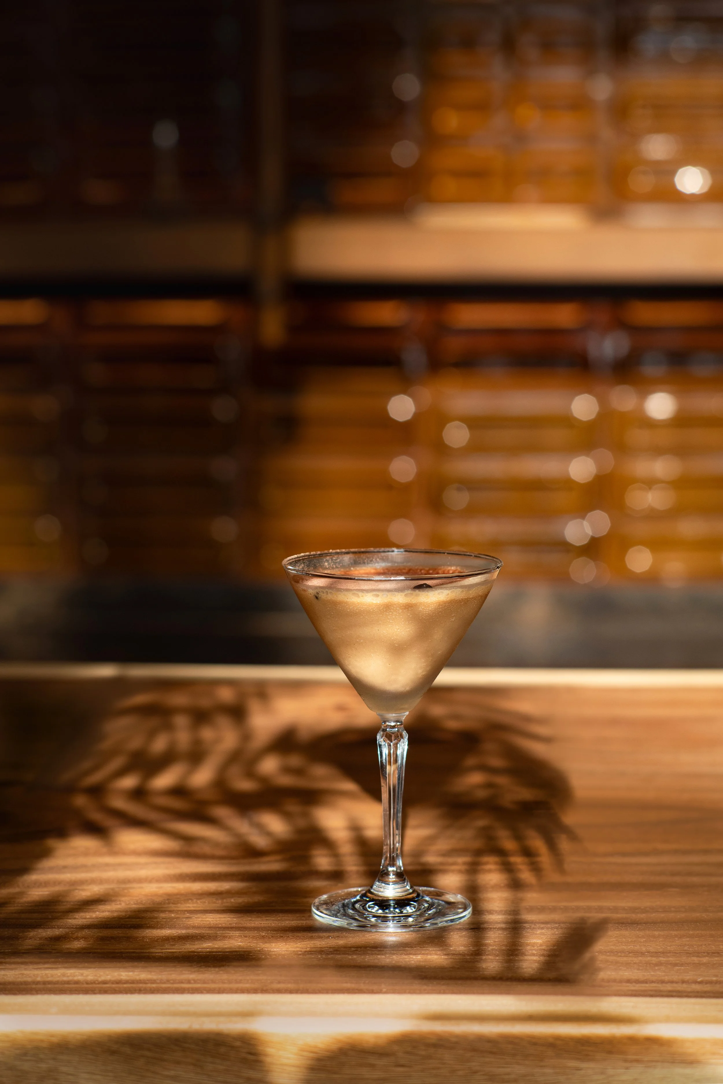 A cocktail in a martini glass with a light-colored liquid and an olive garnish, placed on a wooden surface with shadows cast by nearby plants.