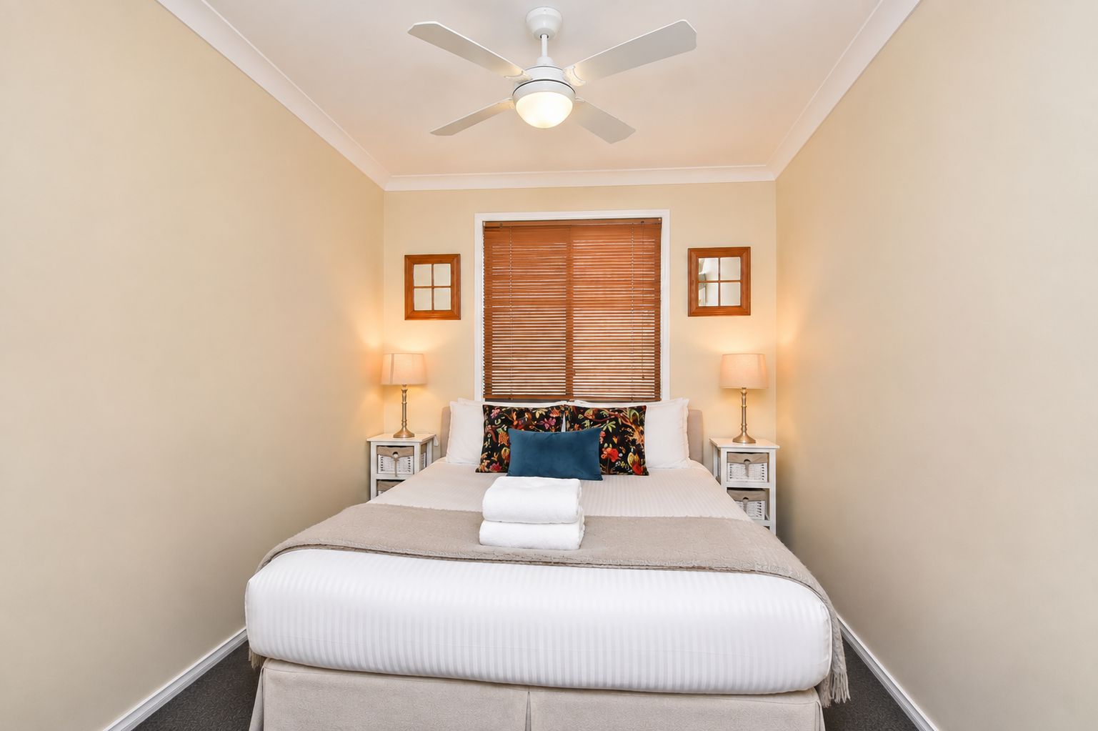 A neatly made bed with white linens, beige blanket, and rolled towels on top. Two nightstands with lamps and decorative pillows in front of a window with wooden blinds in a cozy bedroom.