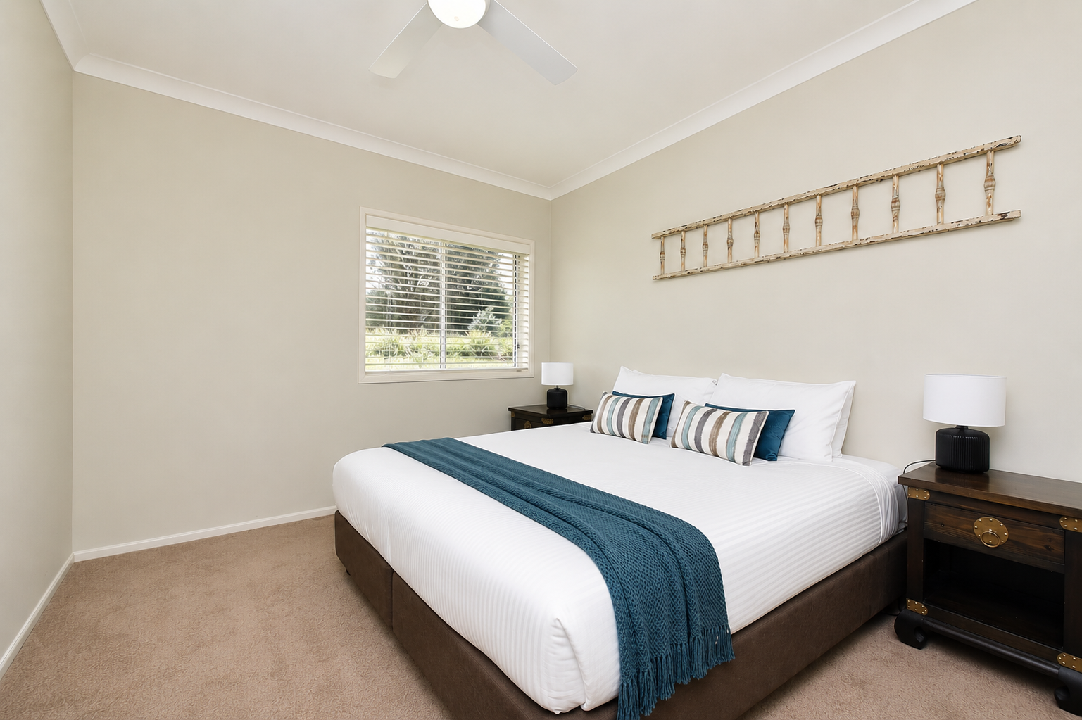 A bedroom with a large bed, white linens, striped and solid pillows, a blue throw blanket, and two dark wood nightstands with lamps. There's a window with blinds and a decorative wooden ladder on the wall.
