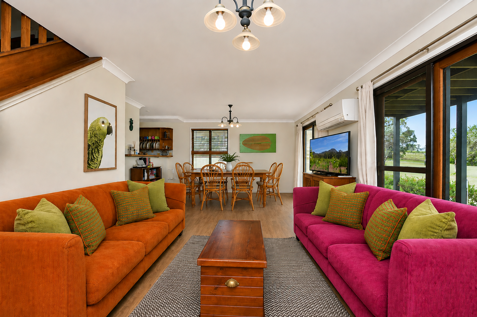 Living room with two colorful sofas, one orange and one pink, with green and patterned throw pillows. A wooden coffee table sits between them on a rug. There is a large window with curtains showing a green outdoor landscape. A flat-screen TV is mount
