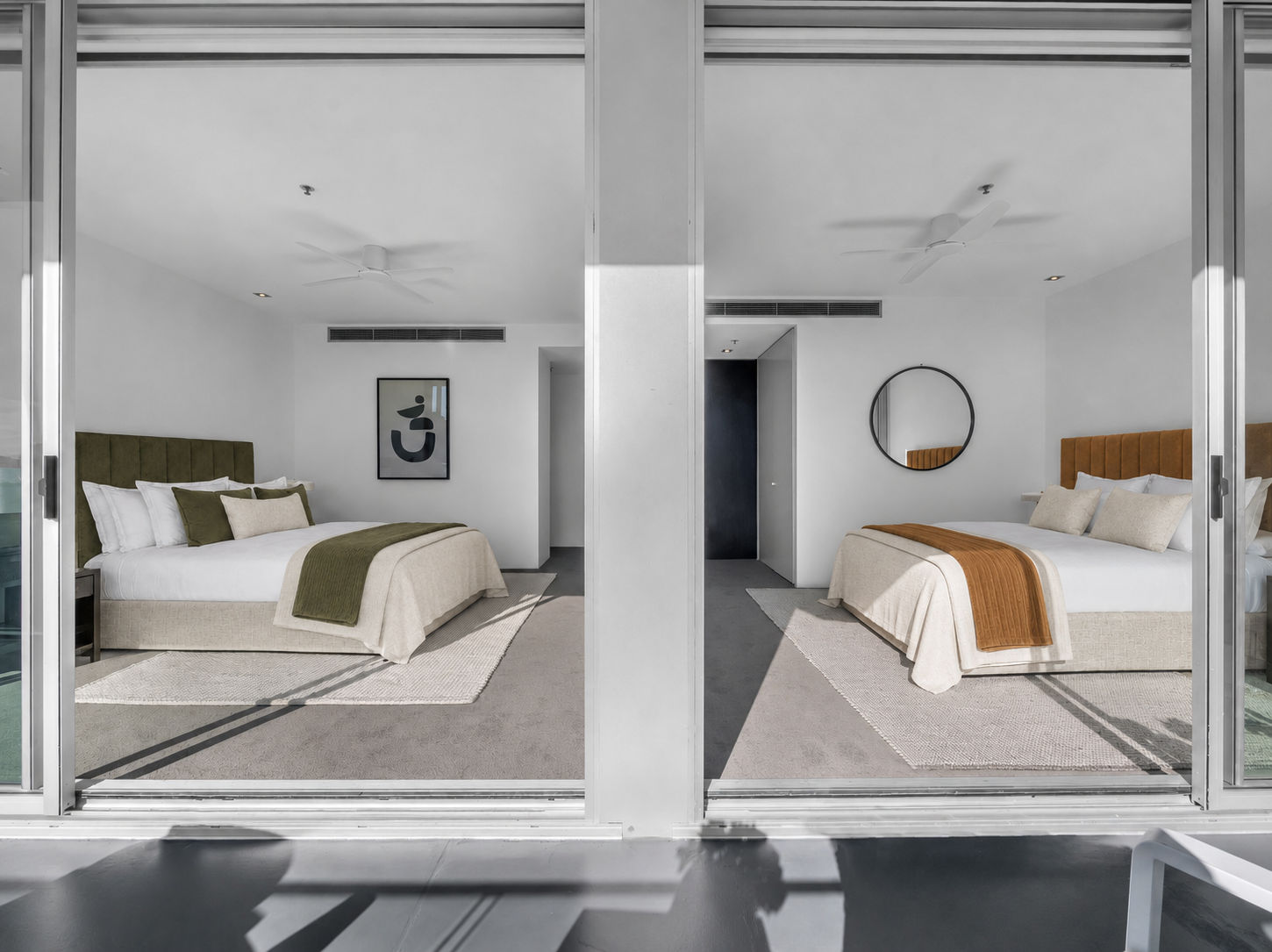 A view through a sliding glass door of two adjacent bedrooms, each with a bed, framed artwork or mirror above, and a carpeted floor.