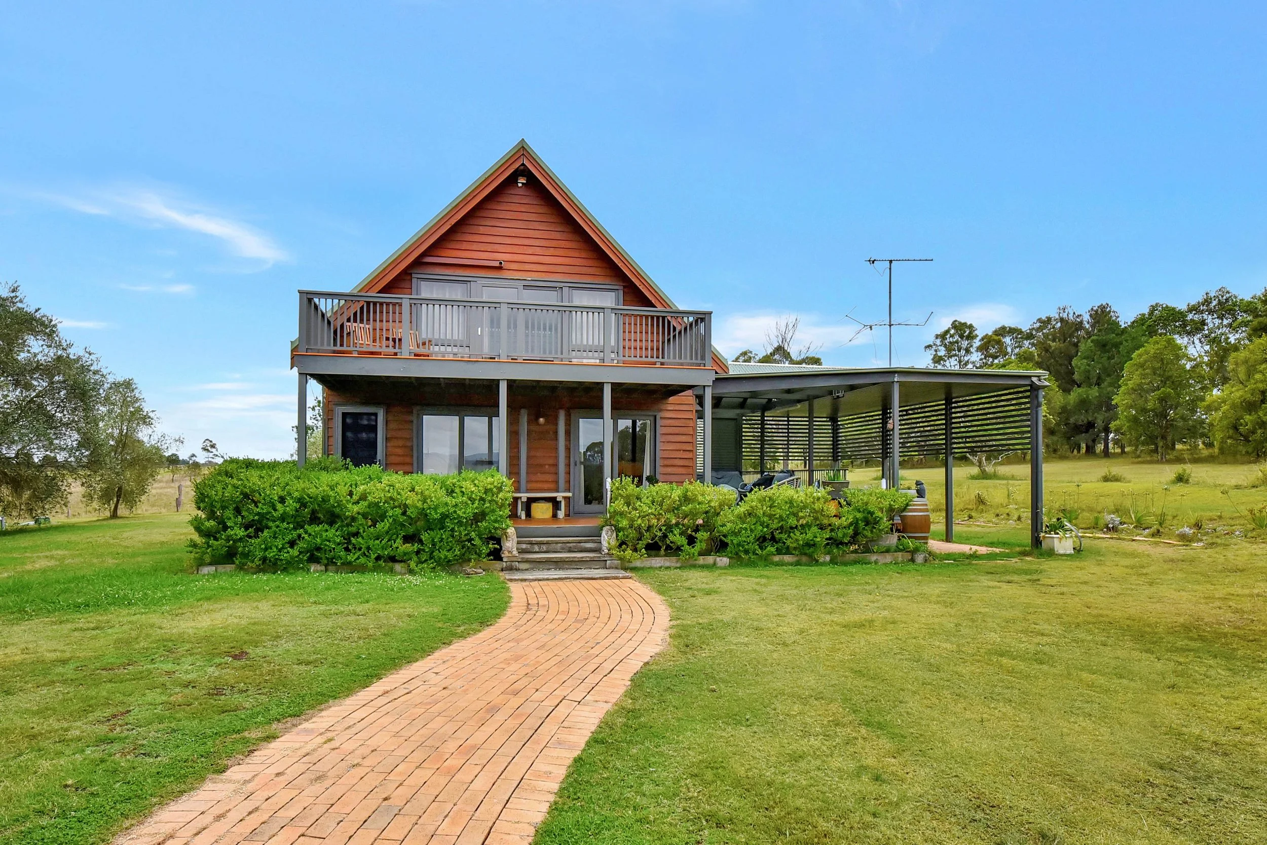 A two-story house made of wood with a balcony and sliding glass doors. There is a brick pathway leading to the house, surrounded by green lawns and bushes. The house has a sloped roof and is set in a rural area with trees and open fields in the backg