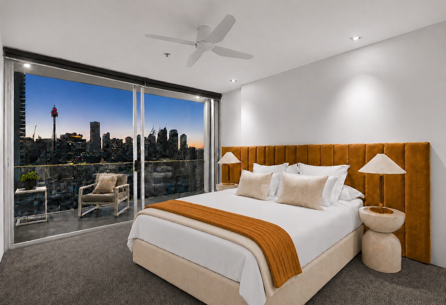 Modern hotel bedroom with large window view of city skyline at sunset, featuring a double bed with orange headboard and white linens, bedside lamps, and a balcony with seating and potted plant.