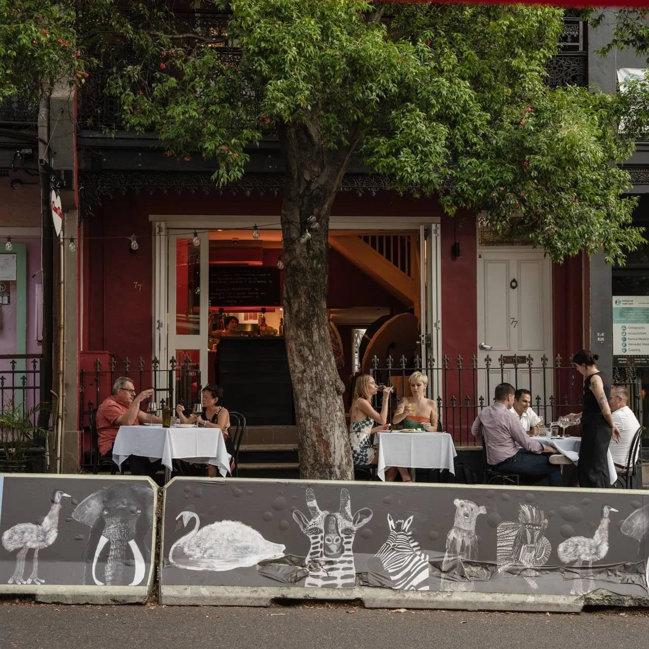 People dining at outdoor tables in front of a restaurant with a large tree and colorful art on the sidewalk barrier.