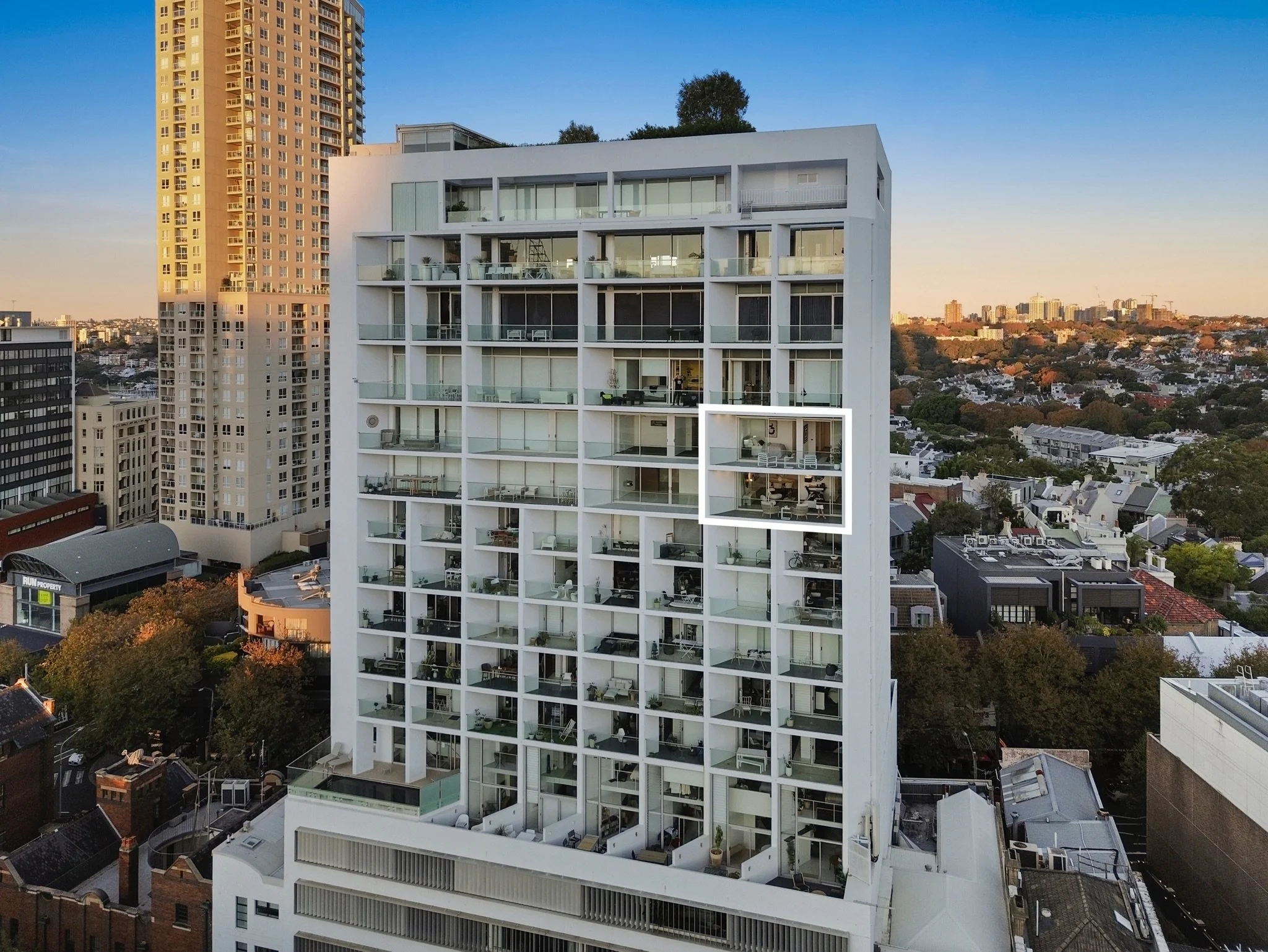 A modern white high-rise apartment building with glass balconies, surrounded by other tall buildings and a cityscape in the background during sunset.
