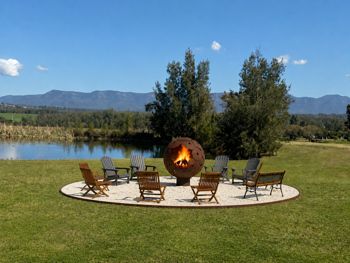 A circle of wooden and metal chairs surrounds a modern firepit with a spherical design, set on a gravel area in a grassy field near a lake, with trees and mountains in the background on a clear, sunny day.
