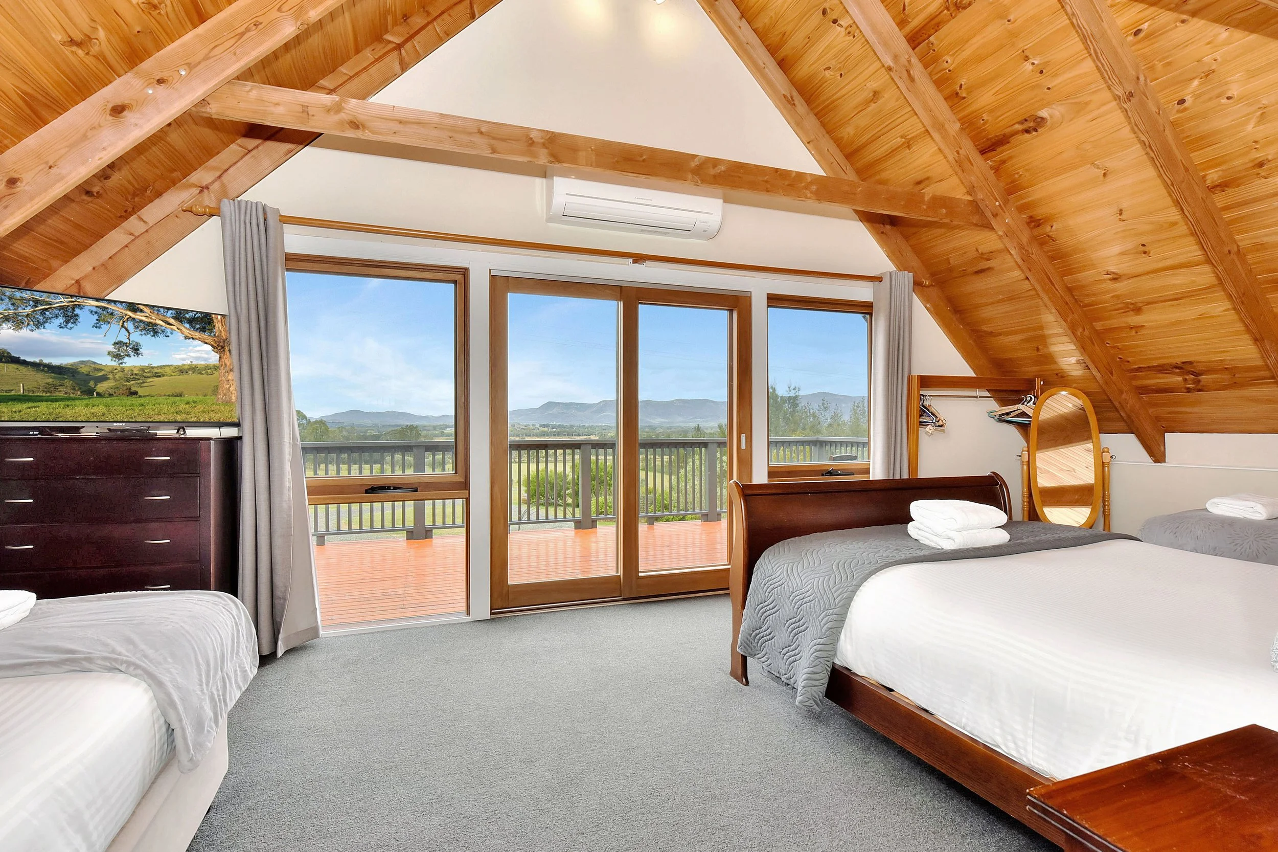 A bedroom with two beds, large windows showing scenic hills and trees, wooden ceiling beams, and a sliding glass door leading to a balcony.