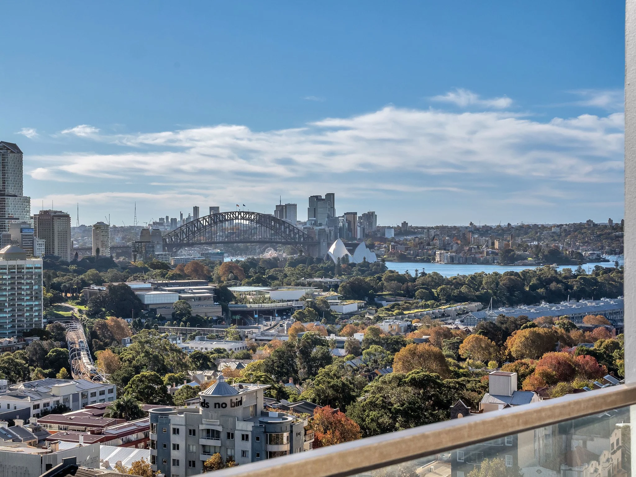 City skyline view with tall buildings, a large bridge, a river, and trees in foreground, under a partly cloudy sky.