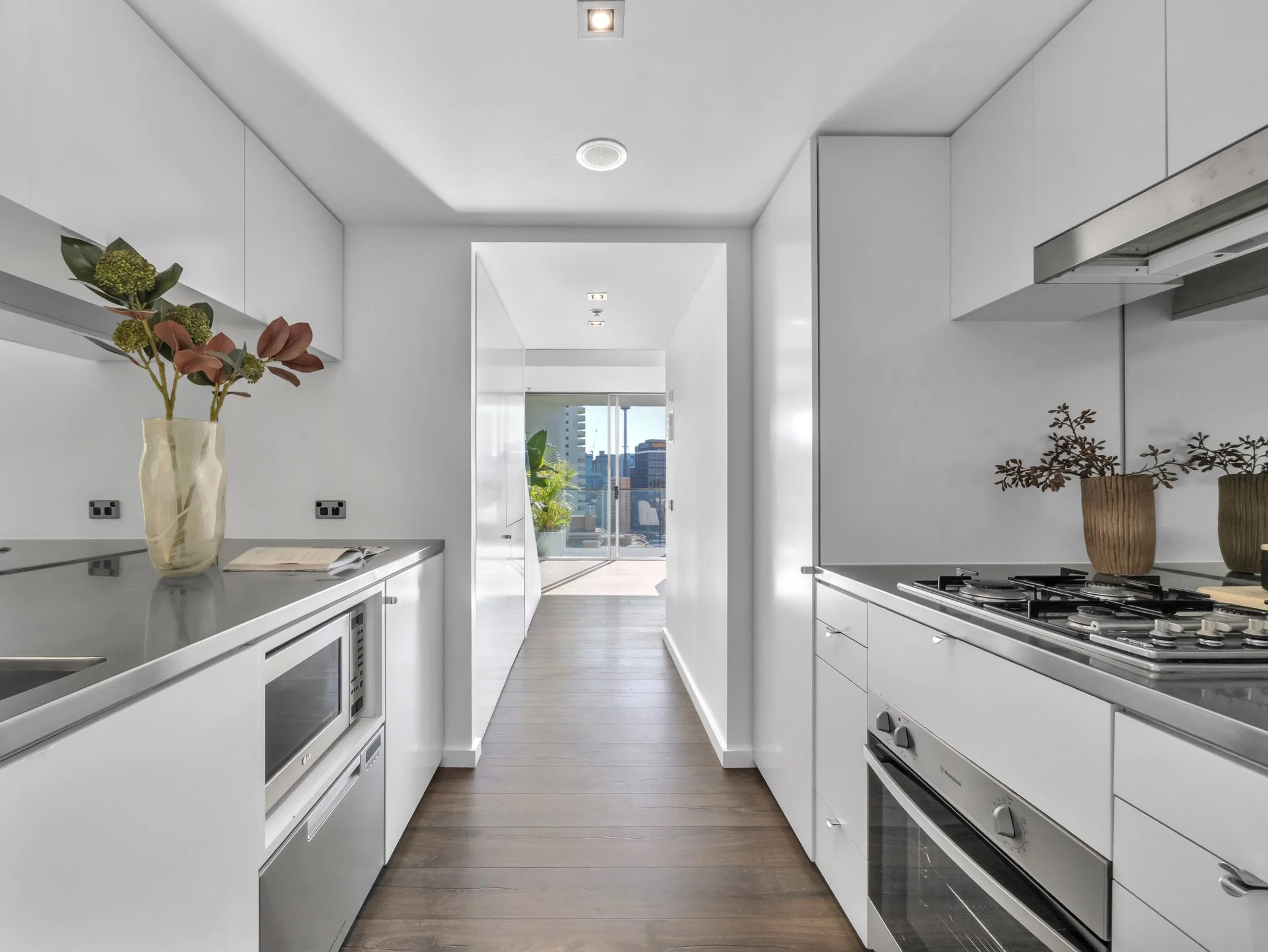 Modern white kitchen with wooden floor, plants on the countertops, city skyline view through the large window, and ceiling lights.