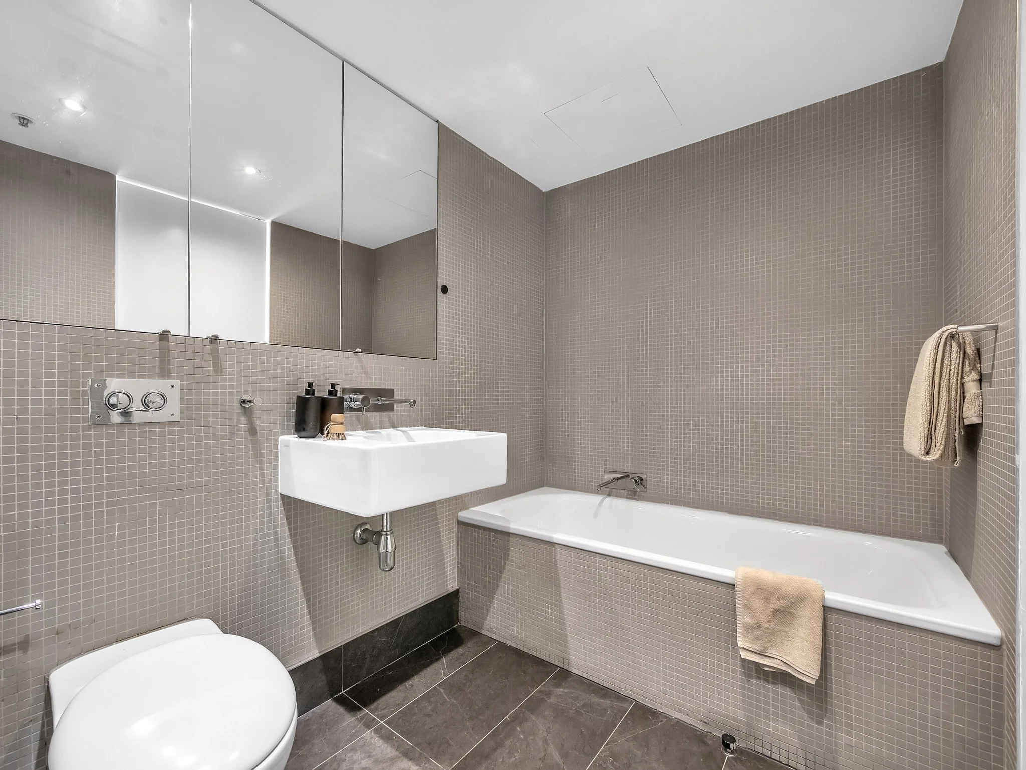 Modern bathroom with beige tile walls, a wall-mounted sink with black soap dispenser, large mirrors, bathtub on the right, toilet in the foreground, and towel hanging on a towel rack.