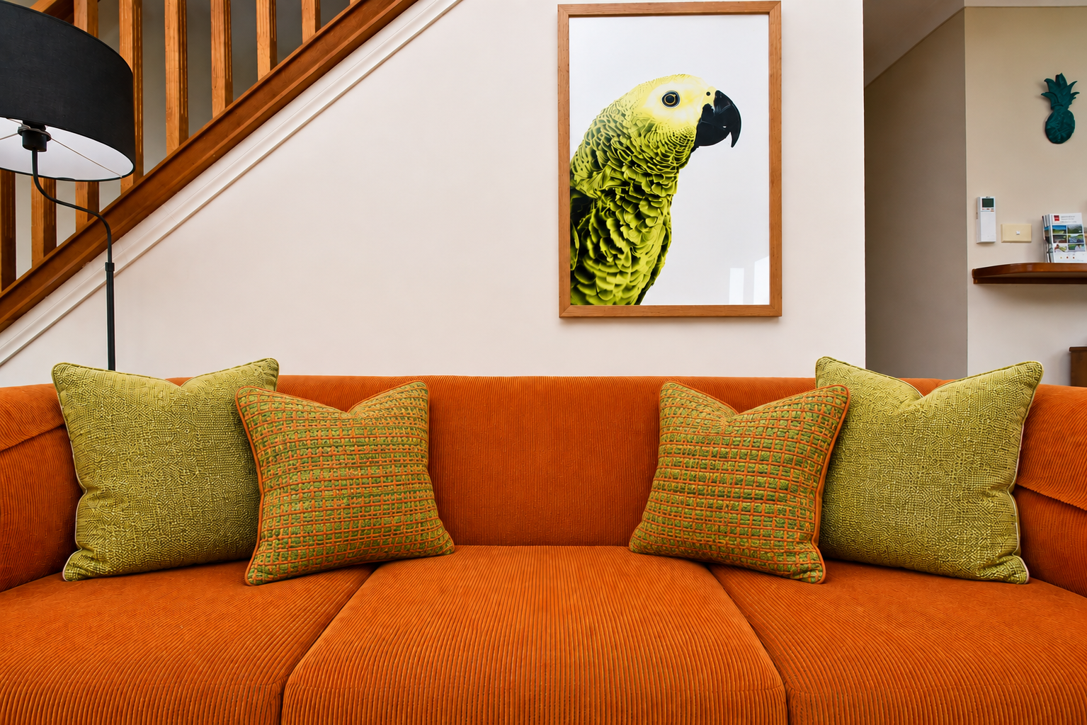 Living room with orange sofa and four green cushions, framed picture of a yellow parakeet on the wall, wooden staircase with railing, and a black floor lamp with a white shade.