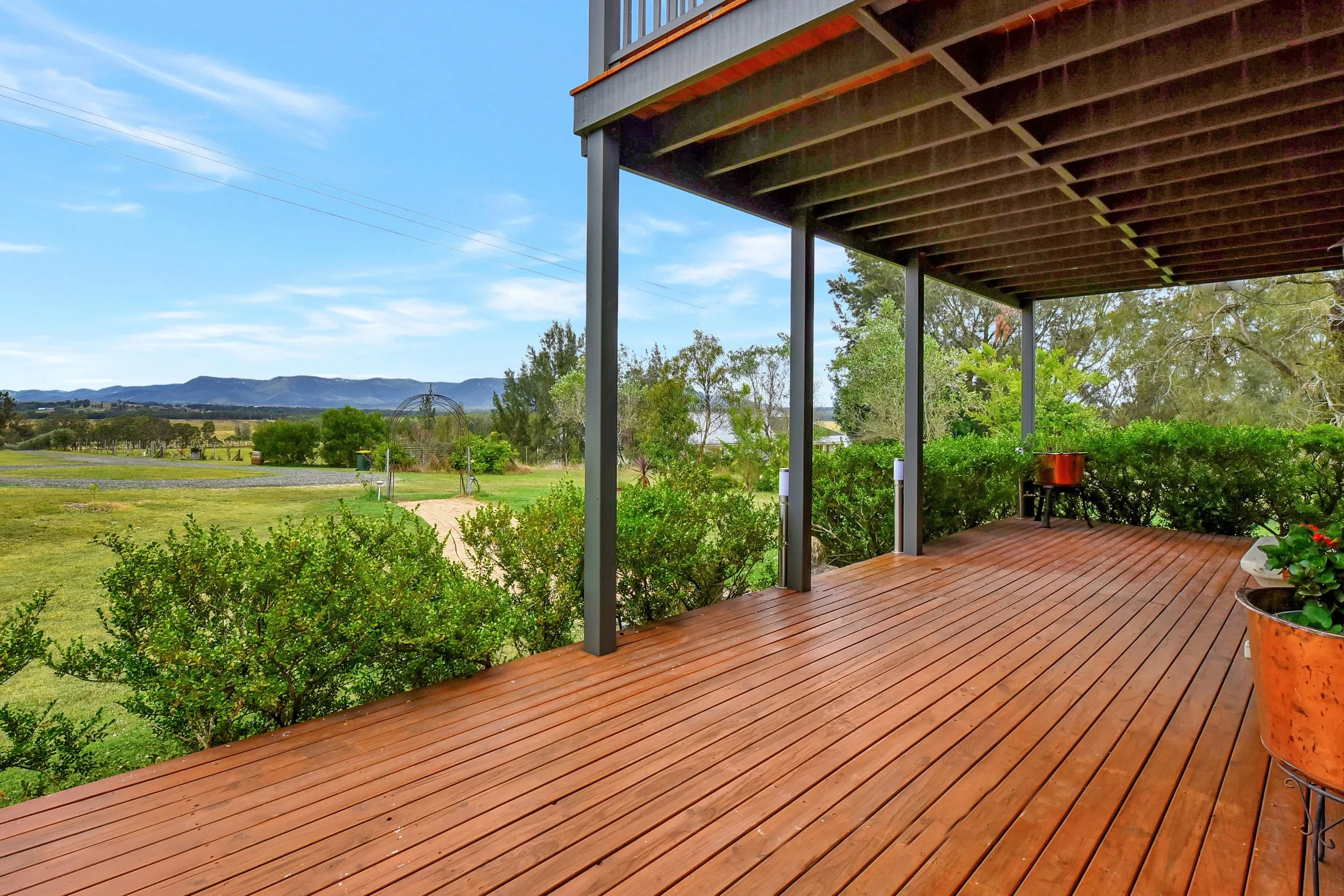 View of a wooden deck porch with flower pots and greenery, overlooking a green field with trees and distant mountains under a blue sky.