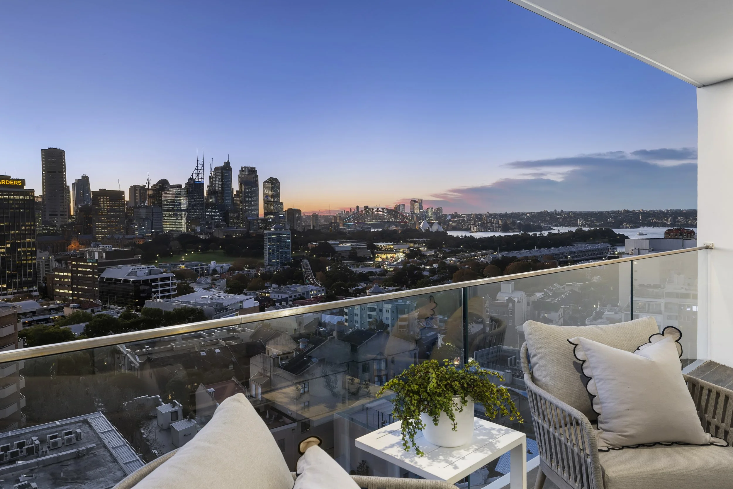 A balcony view of downtown city skyline at sunset, with high-rise buildings, bridge, and river, seen from an outdoor seating area with pillows and a potted plant.
