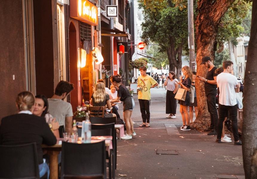 People sitting and standing along a city sidewalk outside a restaurant, with trees and buildings in the background during early evening.
